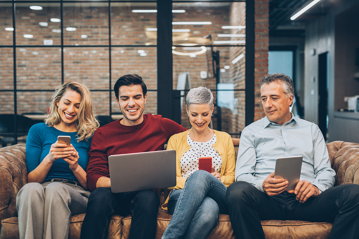 a group of people sitting on a brown leather couch smiling while watching their phones, tablet, and laptop