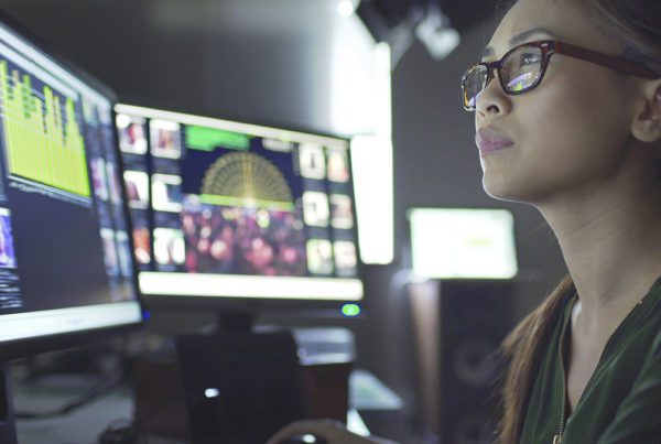 Close up stock image of a young asian woman sitting down at her desk where she’s surrounded by 3 large computer monitors displaying out of focus images of people as thumbnails