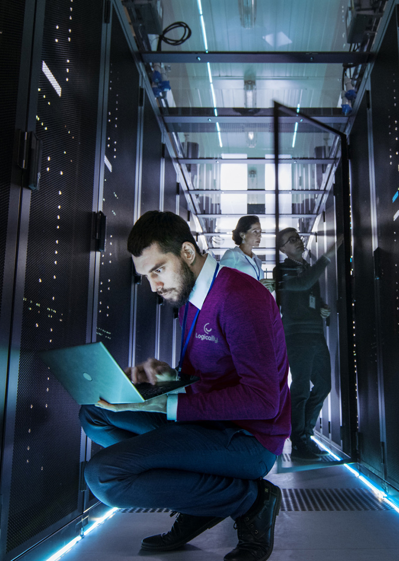 a man wearing a purple logically sweater with a white shirt inside squatting down while holding a laptop with a woman and a man in the background checking a server in a server room