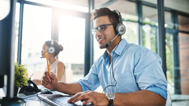 a shot of a male employee working with a headphone on while holding a pencil in front of a computer smiling with a female employee out of focus in the background